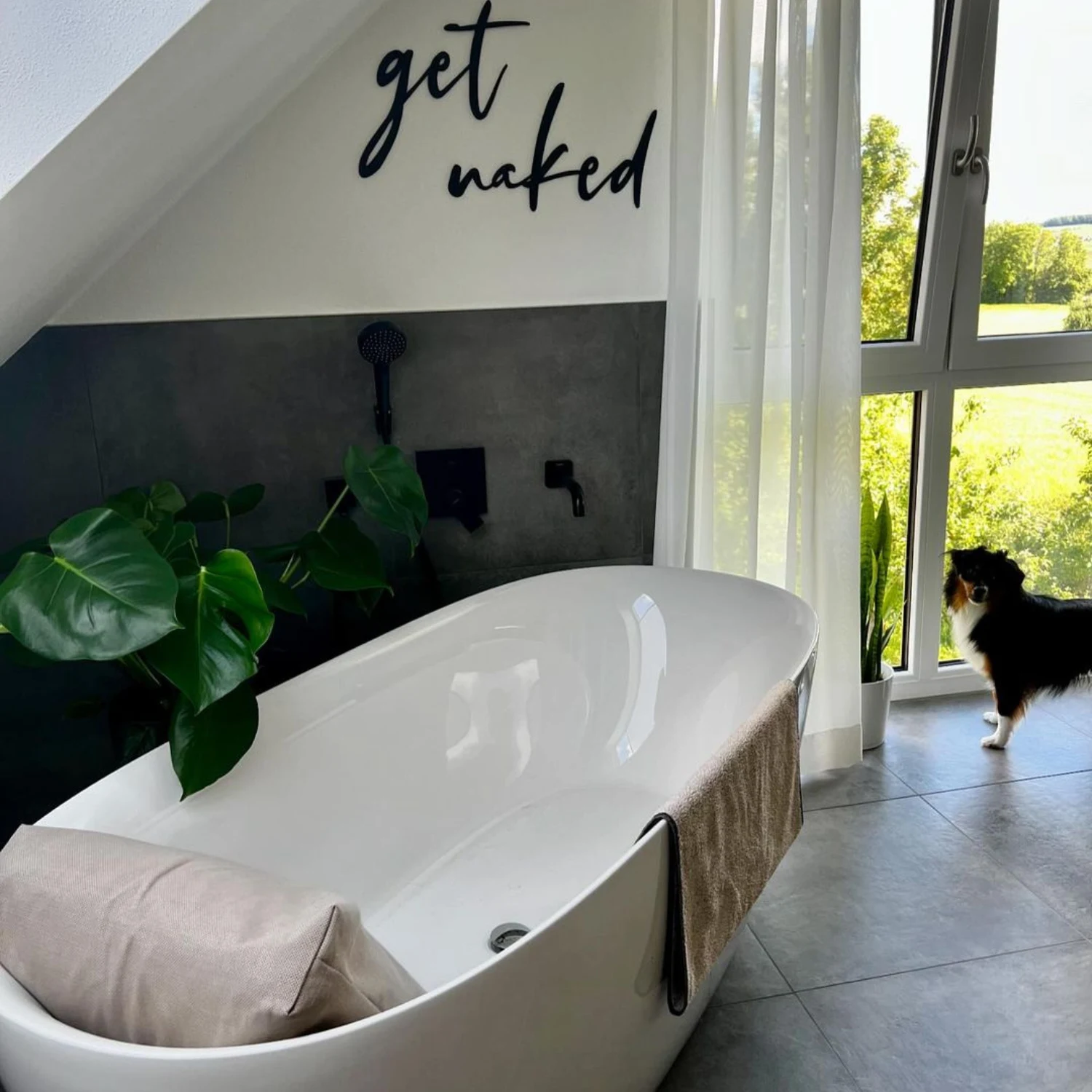 weiße, freistehende Badewanne in einem grau gefliesten Bad mit schwarzen Armaturen und einem schwarzen Schriftzug "get naked" an der Wand