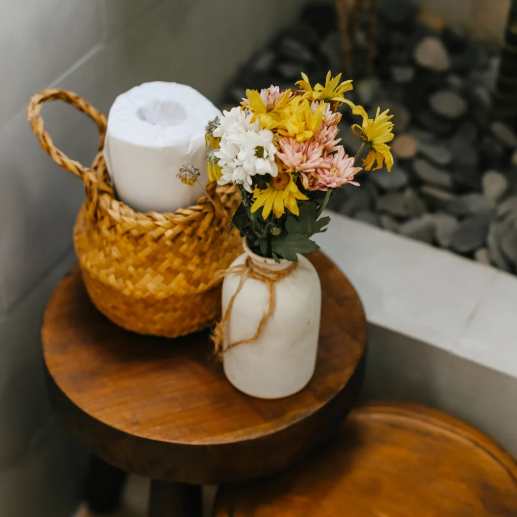 brauner Korb mit Toilettenpapier neben farbfrohen Blumen in einer weißen Vase