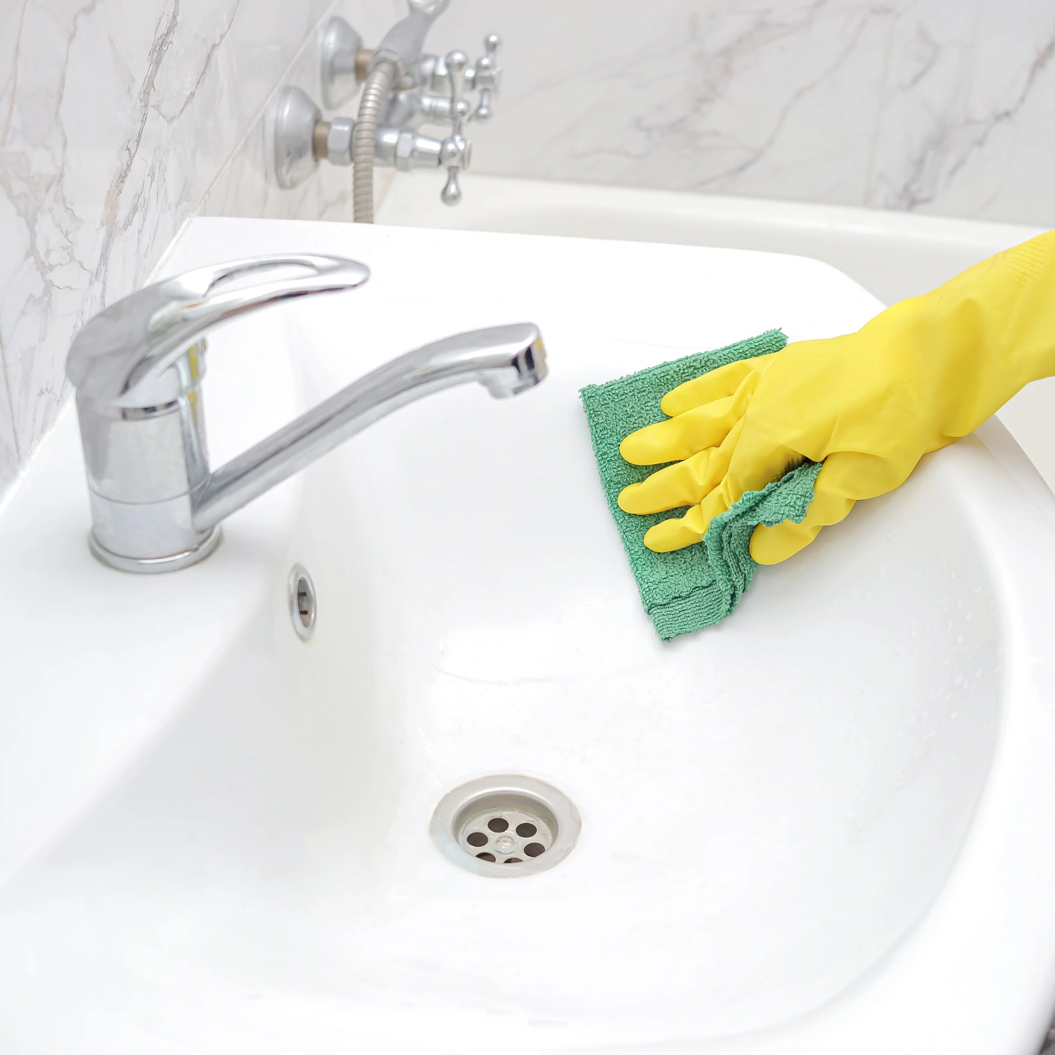 Eine Hand mit gelben Gummihandschuhen säubert das Waschbecken im Badezimmer sorgfältig mit einem Lappen.