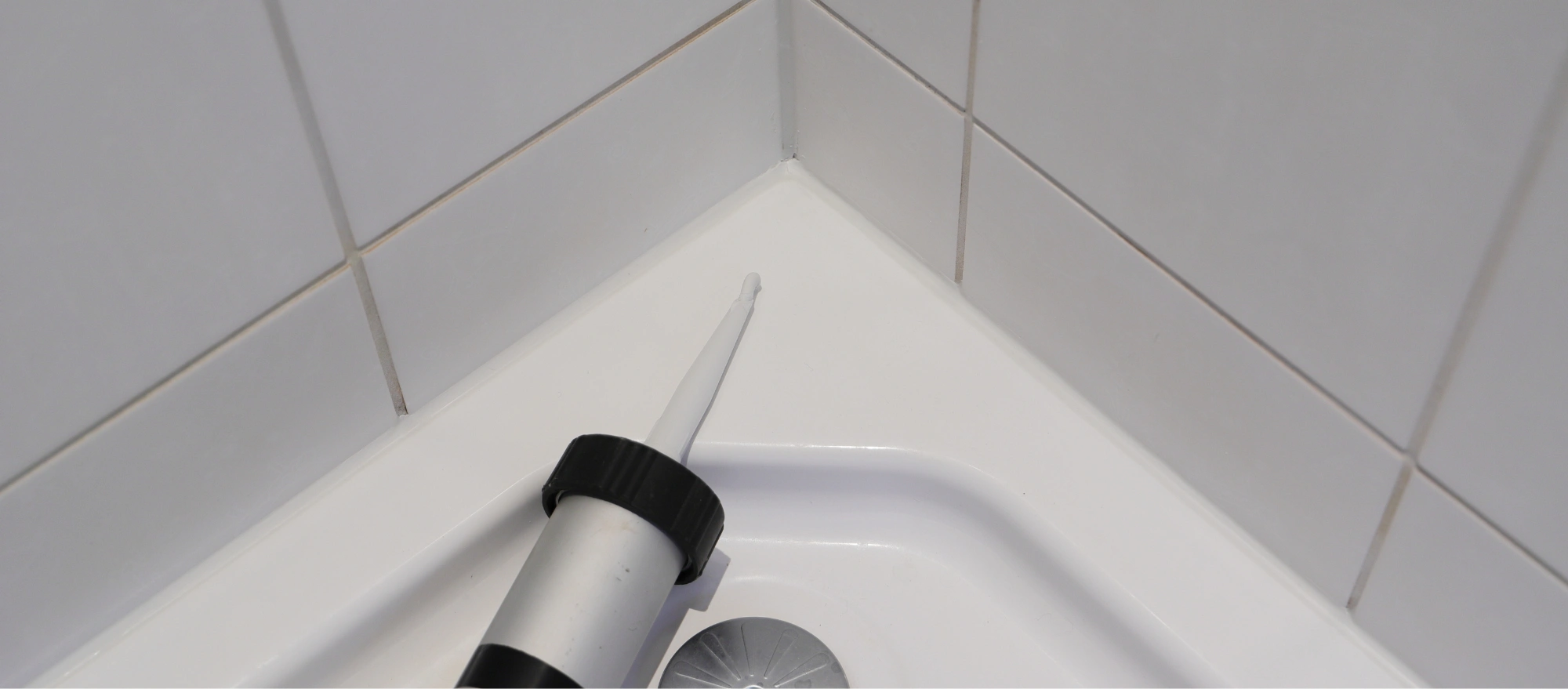 Kartuschenpistole mit weißem Silikon in der Ecke einer Dusche, wo die Fuge zwischen Duschwanne und weißen Wandfliesen abgedichtet wird.
