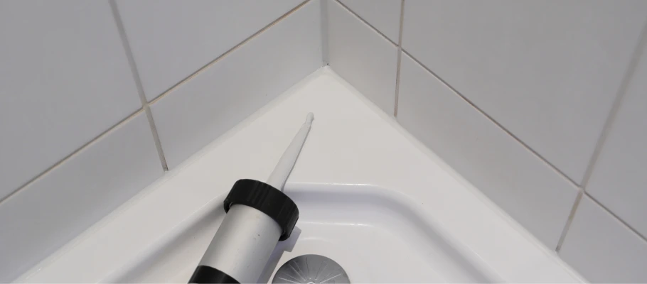 Kartuschenpistole mit weißem Silikon in der Ecke einer Dusche, wo die Fuge zwischen Duschwanne und weißen Wandfliesen abgedichtet wird.